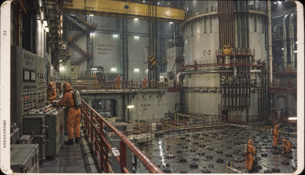 The reactor hall of the atomic pile: a tall cylindrical pile under travelling cranes, technicians in orange protective suits at the control desks and on the chequered floor