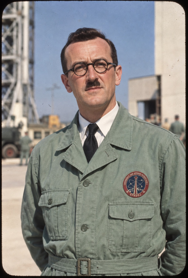 Colour portrait of General Baxter at Sprodj in 1952, wearing pale green Centre overalls with the atomic research badge, launch infrastructure behind him