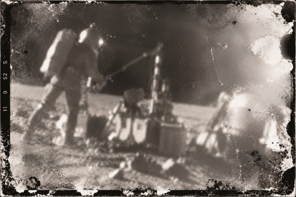Damaged film frame: Frank Wolff, seen from behind in his space suit, setting up equipment on the lunar ground, crates and a striped mast beside him