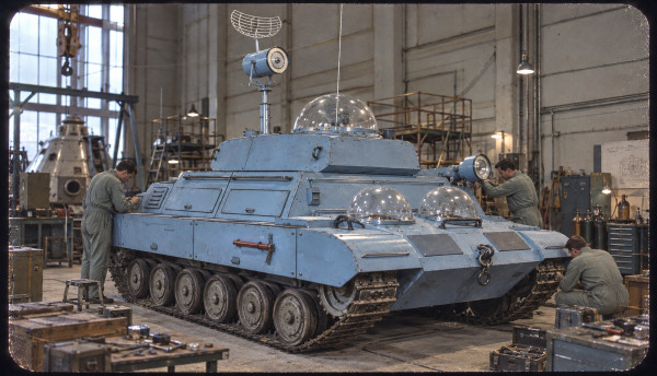 The Moon tank in a workshop at Sprodj: a pale blue tracked vehicle with three glass observation domes, a dish antenna and searchlights on the hull, three technicians in overalls working on it, tool crates and gas cylinders around the bay and a large conical metal section on a stand behind