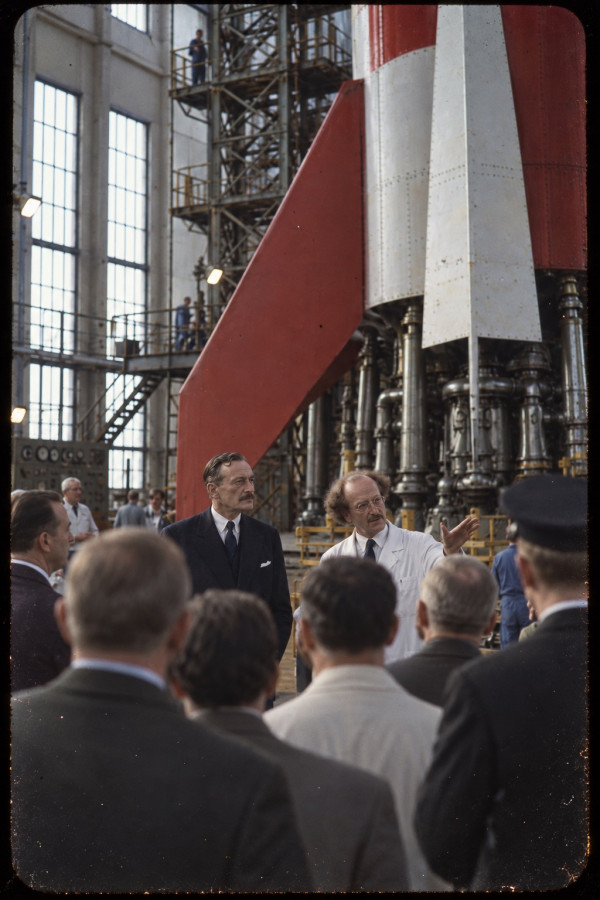 Colour transparency of Muskar XII in a dark suit beside Professor Calculus in a white coat, who gestures towards the red-and-white chequered fin and the cluster of motors of an early rocket in the assembly hall, officials seen from behind in the foreground