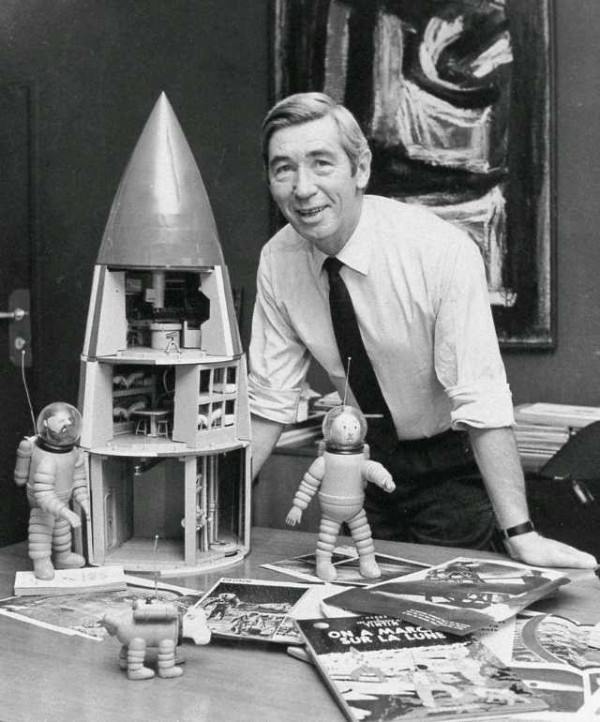 Hergé leaning over a table beside a cut-away model of the Moon rocket, with spacesuited figurines and album plates