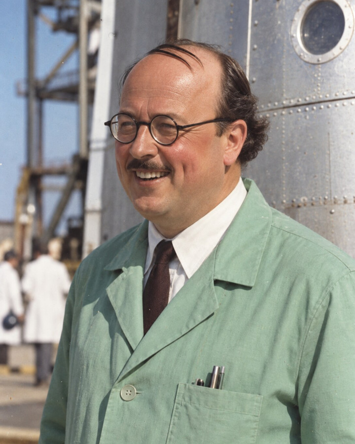 Colour portrait of Frank Wolff, smiling, with round spectacles and a moustache, in a green work coat before a riveted metal hull with a porthole, colleagues in white coats behind