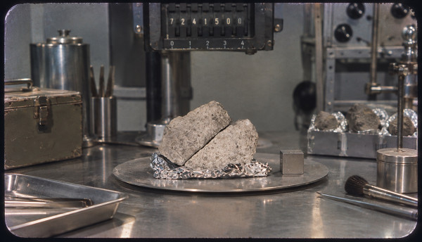 A pale vesicular rock split in two, resting on foil in a steel pan beneath a laboratory balance whose counter reads 7241500, a metal scale cube beside it, more foil-wrapped fragments in a tray to the right, tweezers, a brush and a specimen case on the bench