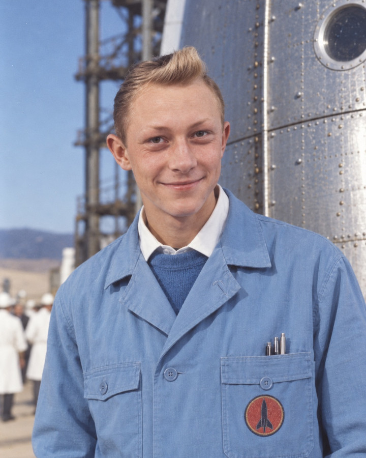 Colour portrait of Tintin, smiling, with his fair quiff, in a blue work coat with a rocket badge on the pocket, before a riveted metal hull with a porthole and a launch gantry, colleagues in white coats behind