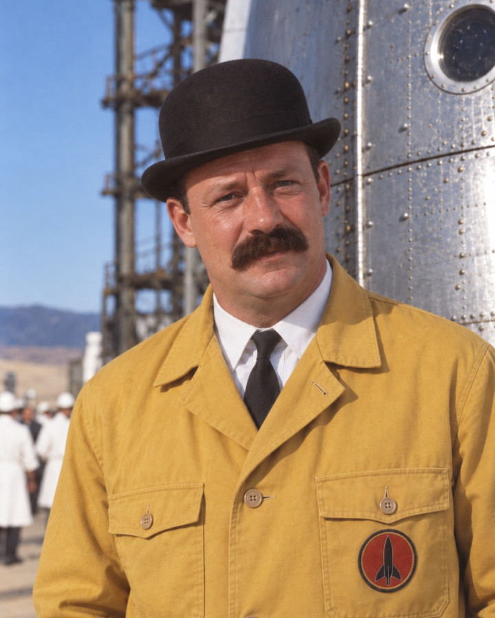 Colour portrait of Thompson, in a black bowler hat and a mustard-yellow work jacket with a red rocket badge on the pocket, white shirt and black tie, his moustache cut straight, before a riveted metal hull with a porthole and a launch gantry, staff in white coats behind