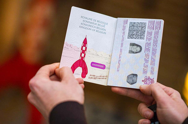 Two hands holding an open Belgian passport: the left page carries the Kingdom's name in French, Dutch, German and English above the red-and-white chequered rocket standing on a pale mountain landscape, with the specimen number GA0000000; the right page is the data page, with its photograph, barcode and security printing