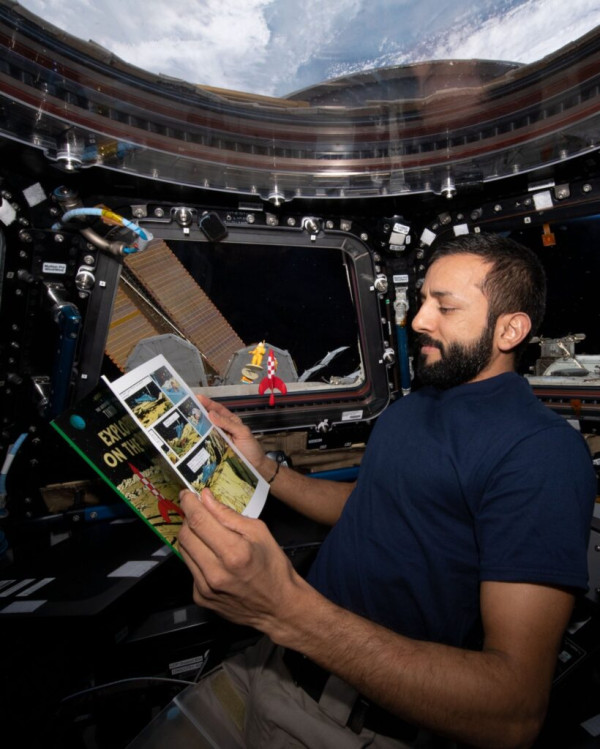 Photograph inside the space station's cupola: an astronaut in a dark polo shirt holds an open comic album with both hands, reading; behind him, by the window, a small figure in an orange spacesuit and a red-and-white chequered rocket float in mid-air, with the station's structure and the Earth's limb beyond