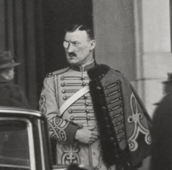 Black-and-white photograph of Colonel Boris Jorgen standing in a palace doorway, a monocle at his eye, in a frogged hussar-style dress tunic with a fur-trimmed pelisse over one shoulder and a white crossbelt across his chest; a motor car and onlookers at the edges of the frame