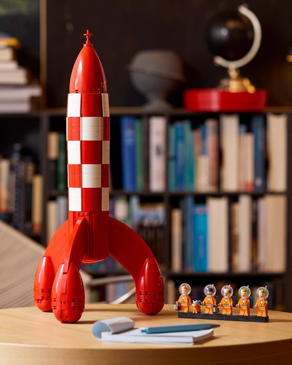 Photograph of a brick-built model of the chequered red-and-white rocket on a wooden table, five small figures in orange spacesuits and a white dog on a base beside it, a bookcase and a globe out of focus behind, a notebook and pen in front