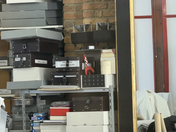 A small red-and-white chequered model of the Moon rocket stands on a shelf among dark storage boxes with leather straps, one labelled PHARMACIE MONSIEUR ALAÏA, in front of a brick wall; other boxes are labelled CRAIE, and the gilded frame of a tall mirror stands to the right