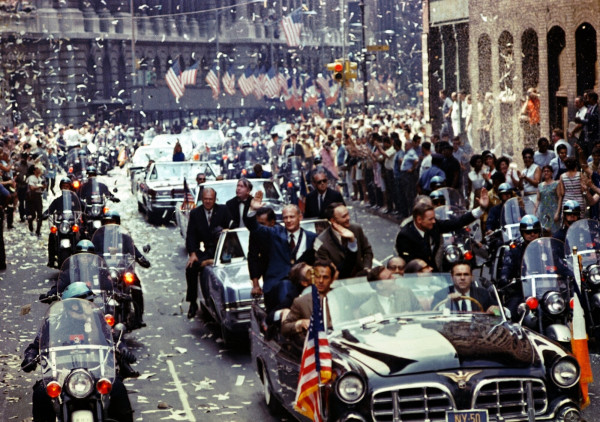 Colour photograph of the Apollo 11 astronauts waving from an open car on Broadway amid falling ticker tape, escorted by police motorcycles, crowds and American flags on both sides of the street