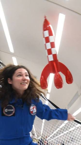 Photograph inside an aircraft cabin: a woman in a blue flight suit with agency patches, her hair floating, looks up at a red-and-white chequered toy rocket drifting weightless below the ceiling lights, safety netting to the right