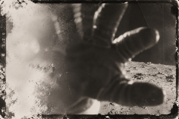 Damaged film frame: a gloved hand fills the foreground above the lunar surface, the exposure heavily fogged