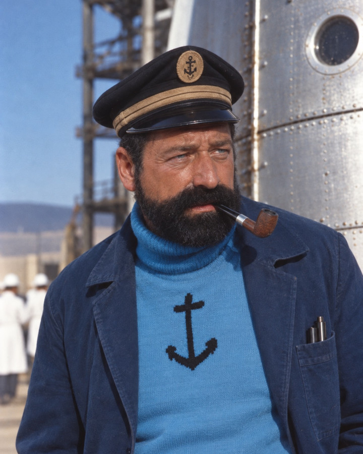 Colour portrait of Captain Haddock, bearded, pipe in mouth, in a peaked captain's cap with an anchor badge, a blue roll-neck sweater bearing a black anchor and a navy work jacket, looking aside, before a riveted metal hull with a porthole and a launch gantry