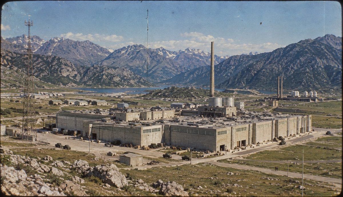 Aerial colour view of the Sprodj Atomic Research Centre: long concrete halls, a tall chimney and a radio mast in a wide valley ringed by snow-capped mountains
