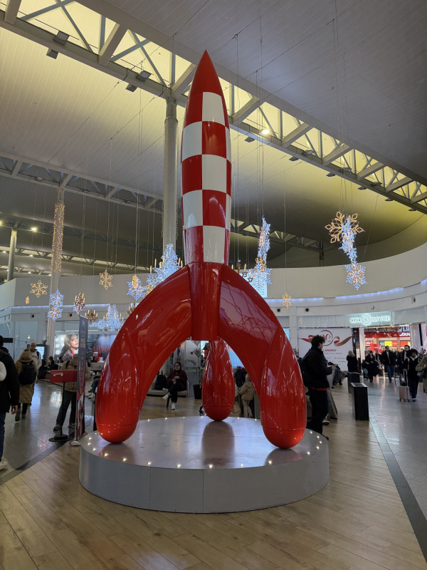 A tall red-and-white chequered rocket sculpture standing on a lit circular plinth in an airport concourse, illuminated snowflake decorations hanging from the roof beams, duty-free shops and travellers with luggage around it