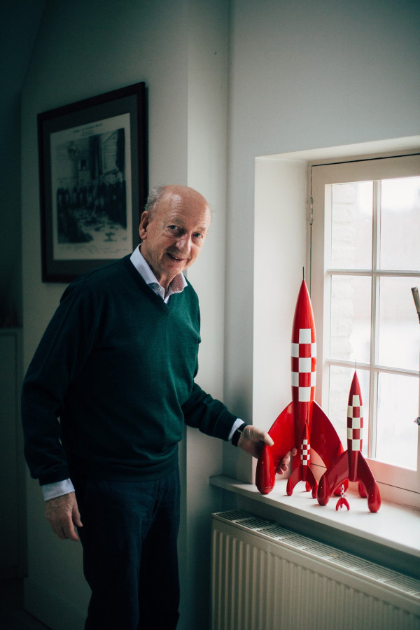 Dirk Frimout, in a green V-neck jumper over a light shirt, smiles beside a window, one hand on the largest of several red-and-white chequered models of the Moon rocket lined up on the sill; a framed black-and-white print hangs on the wall behind him