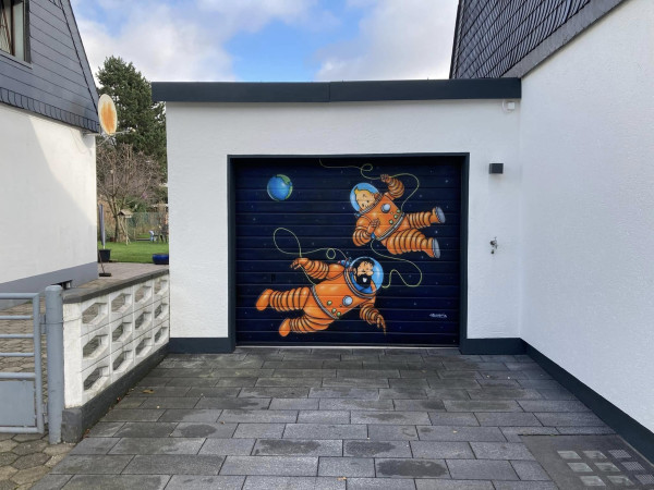 A white garage between two slate-roofed houses, reached by a paved driveway: its door is painted with a mural of Tintin and Captain Haddock in orange spacesuits floating among the stars, each holding a loose yellow line, with the Earth in the upper left and the painter's signature at lower right