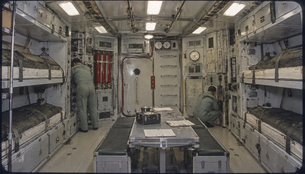 The crew cabin of the lunar rocket: bunks along both walls, a narrow table with papers down the middle, two men in overalls at work