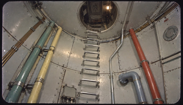 Looking up inside the hold of the lunar rocket: a riveted aluminium shaft with pipe runs colour-coded green, cream, grey and red, a ladder of steel rungs climbing the wall to an open circular hatch with a lit compartment beyond, brass conduit, a junction box and a round inspection port