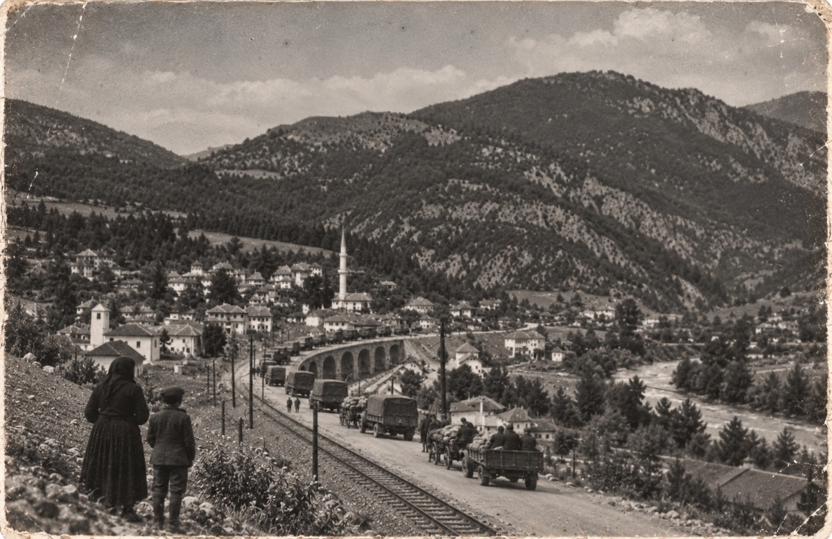 Black and white photograph taken from an embankment above a valley road: a woman in a headscarf and a boy in a cap stand with their backs to the camera, watching a column of canvas-topped lorries and horse carts move through; below them a railway line and a stone viaduct, and beyond, a town of white houses with a minaret and a church bell tower under wooded mountains, a river running along the valley on the right; the print is creased and worn at the corners
