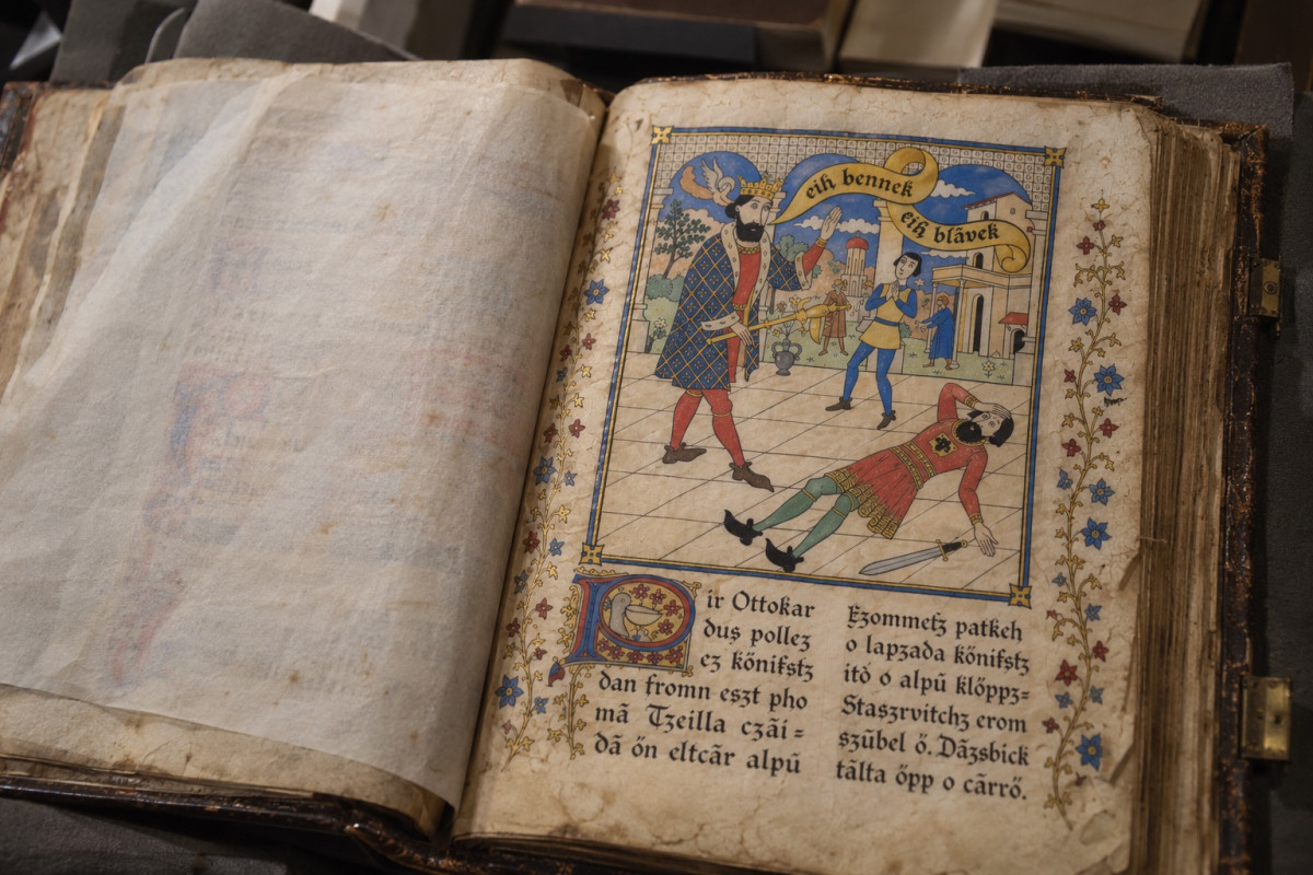 The manuscript lying open on grey foam supports in a library store, seen at an angle, a tissue guard over the left-hand page: on the right, a painted miniature in which the crowned King Ottokar IV holds the sceptre, Baron Staszrvitch lies struck down on a tiled floor with his sword beside him, and a banner reads eih bennek eih blavek; below, lines of Syldavian script framed by painted flowers