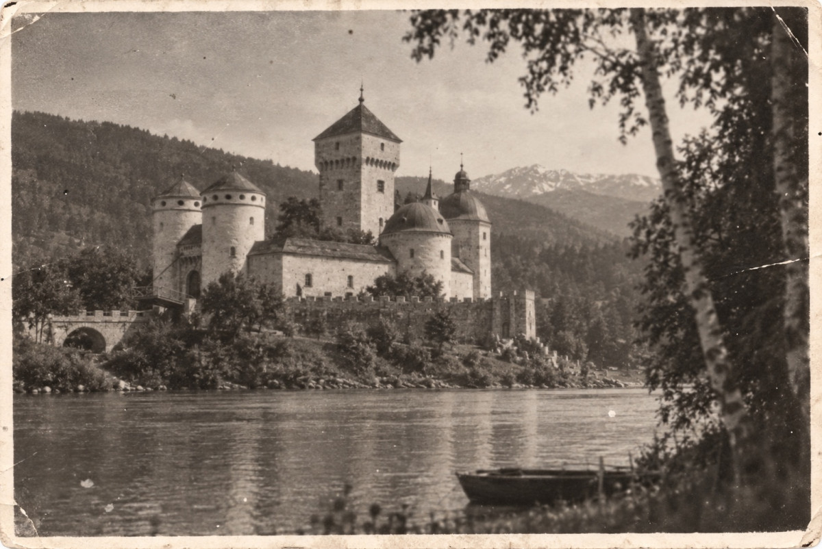 Kropow Castle across a wide river, seen from the far bank through silver birches: the tall square keep, domed towers and round towers in front of it, crenellated ramparts along the water and wooded mountains behind; in the foreground an empty rowing boat lies by the bank