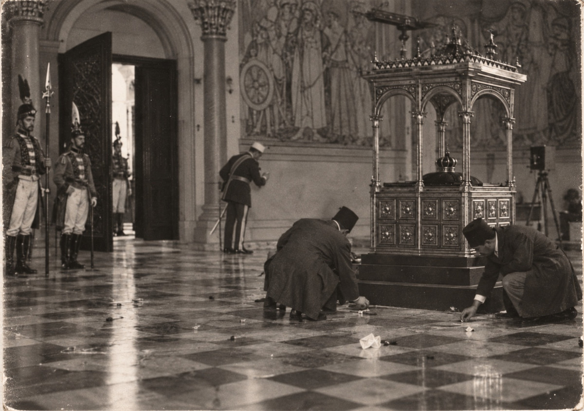 An old photograph of the Treasure Chamber: the gilded case under its canopy with the crown inside, a camera on its tripod against the far wall below large painted figures; two men in fezzes crouch and search the chequered floor among scattered small objects, an officer in a white cap, sword at his side, taps the wall by a column, and guards in plumed caps with halberds stand at the open double door