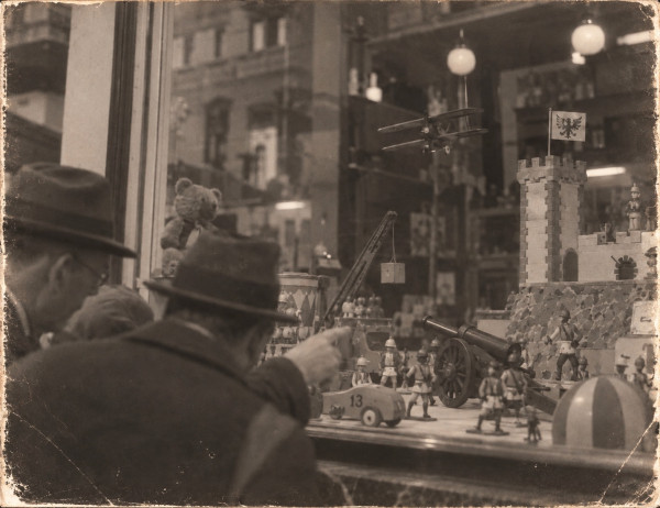 Two men in hats and overcoats at a toy-shop window, seen from behind; one points at a toy cannon on wooden wheels among tin soldiers, beside a toy castle, a racing car numbered 13, a crane, a teddy bear and a model biplane hanging above
