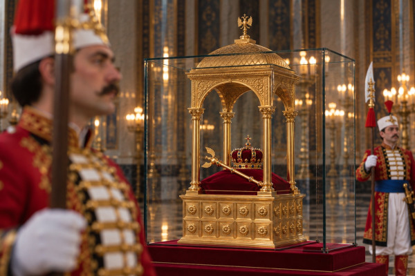 The gilded case in the Treasure Chamber, a canopy on golden columns inside a glass cabinet: the jewelled crown and the Sceptre of Ottokar, its pelican with raised wings, lie on a red velvet cushion; a guard of the Treasure Guard in a red and gold coat stands out of focus in the foreground, halberd in hand, and a second guard with a halberd and a blue sash keeps watch on the right