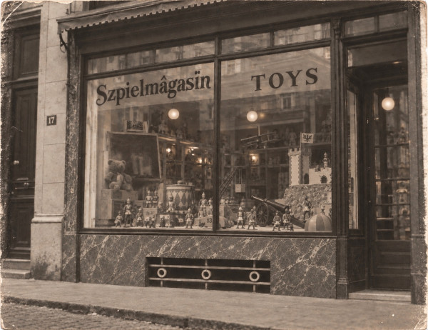 Old photograph of a shop front on a cobbled street: a large window lettered Szpielmâgasin and TOYS, filled with tin soldiers, a toy spring cannon on wooden wheels, a toy castle flying a flag, a racing car numbered 13, a drum, a crane, a ball, a teddy bear and a model biplane; a marble base below the window, the door open on the right, the house number 17 on the wall at left