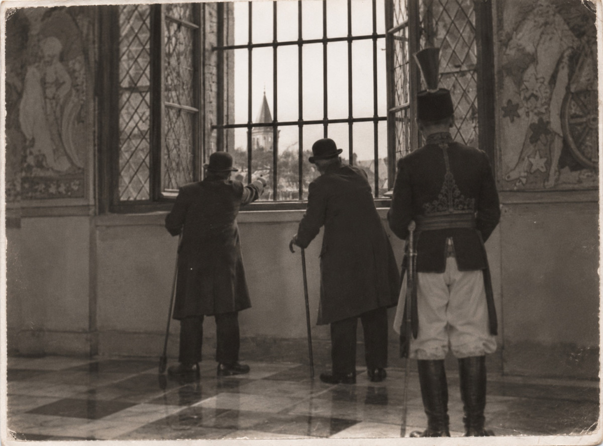 An old photograph taken from behind: two men in bowler hats and long coats, leaning on walking sticks, look out through a tall barred window with its leaded casements open, one pointing outside towards a pointed spire and rooftops; a guard in a tall plumed shako, embroidered jacket, white breeches and boots, sabre at his side, stands behind them; painted figures on the walls either side, a chequered floor
