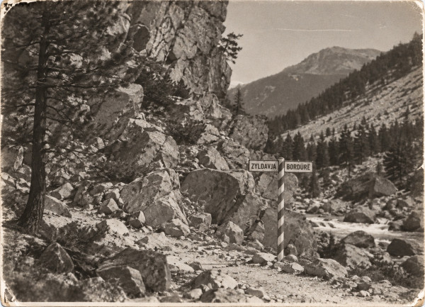 An old photograph of a mountain pass: a striped frontier pole with two signs, ZYLDAVJA and BORDÜRË, stands on a stony track among fallen boulders below a rock face and a pine; a torrent runs on the right, wooded slopes and a bare peak behind