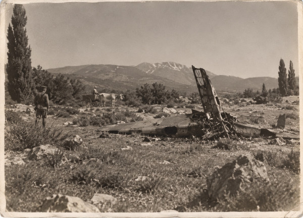 An old photograph of a stony upland meadow: the torn wreck of a monoplane lies flat in the scrub, a wing with its skin ripped open and a tail fin reduced to a bare frame standing upright; a soldier in a cap and puttees looks on from the left, an ox-cart and its driver pass behind, cypresses and a snow-streaked mountain in the distance
