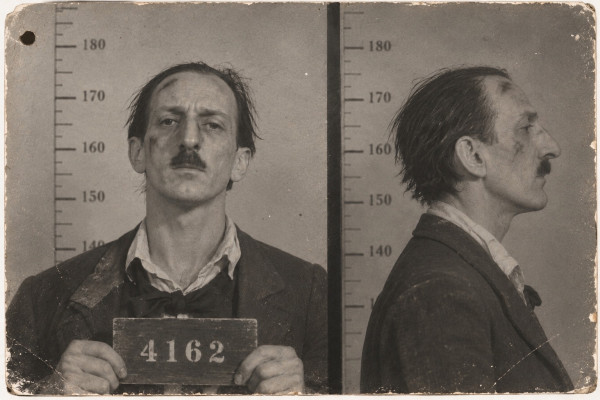 Police identification photograph, full face and profile side by side against a height scale: a thin man with lank, dishevelled hair, a short moustache and smudges of soot on his face, in a crumpled jacket with a loose bow tie, holding a placard numbered 4162; a filing hole in the corner