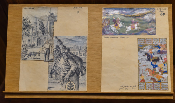 Two documentation sheets on a wooden stand: on the left, one labelled “Albanie” no. 17, with engravings of a street with a mosque and a donkey cart and of a bearded vendor carrying a large brass jug; on the right, one labelled “Iran” no. 54, with a Persian miniature of riders hunting on white horses and a polo game framed by Persian script, each with a handwritten caption