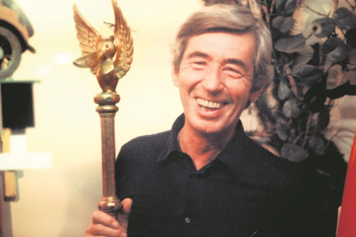 Hergé, smiling, in a dark shirt, holds up a gilded sceptre topped by a pelican with spread wings