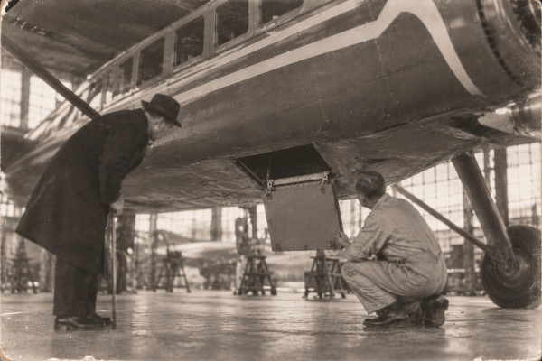 An old photograph in an aircraft hangar: a single-engined monoplane seen from below, a square trapdoor open in the belly of its fuselage; a mechanic in overalls crouches and holds the hinged panel, while a man in a hat and a long dark coat, leaning on a cane, stoops to look into the opening; other aircraft stand on trestles in the background
