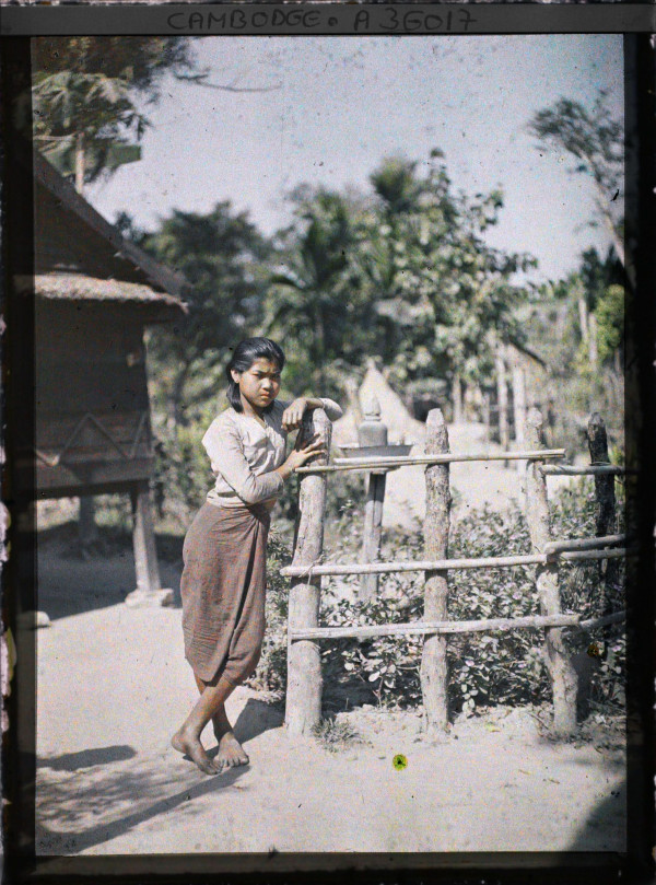 Autochrome of young Cambodian girl leaning on barrier with spirit house visible behind