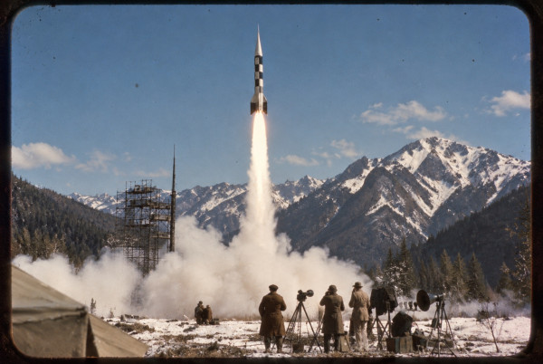 A chequered test rocket lifting off from its scaffold gantry at the Sprodj range, newsreel cameramen at their tripods in the snow in the foreground, snow-covered mountains behind, 1950